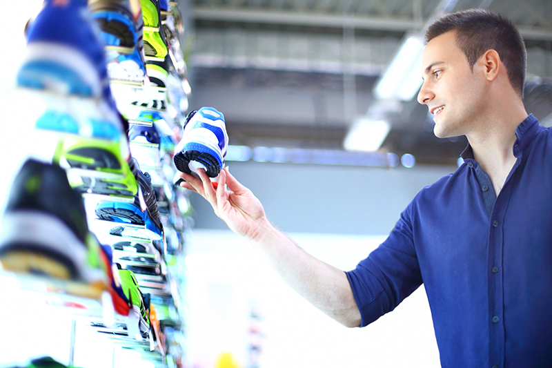 Young caucasian man in shoes store buying a jogging pair. He's holding one shoe and looking at it, smirk of satisfaction shows up on his face. He has short brown hair and navy blue shirt with sleeves rolled back